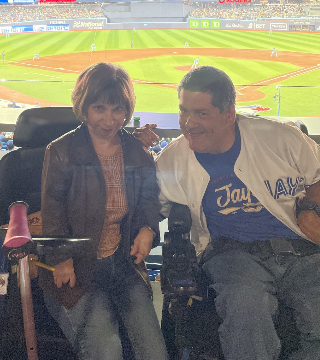 A man and woman sit next to each other in the stands of a baseball stadium, with the baseball diamond visible in the background.