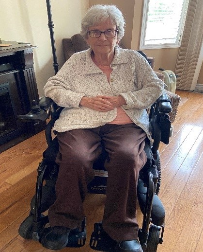 Denise, who has short, white-grey hair and glasses, and wears a white cardigan and brown pants, sits in a living room.