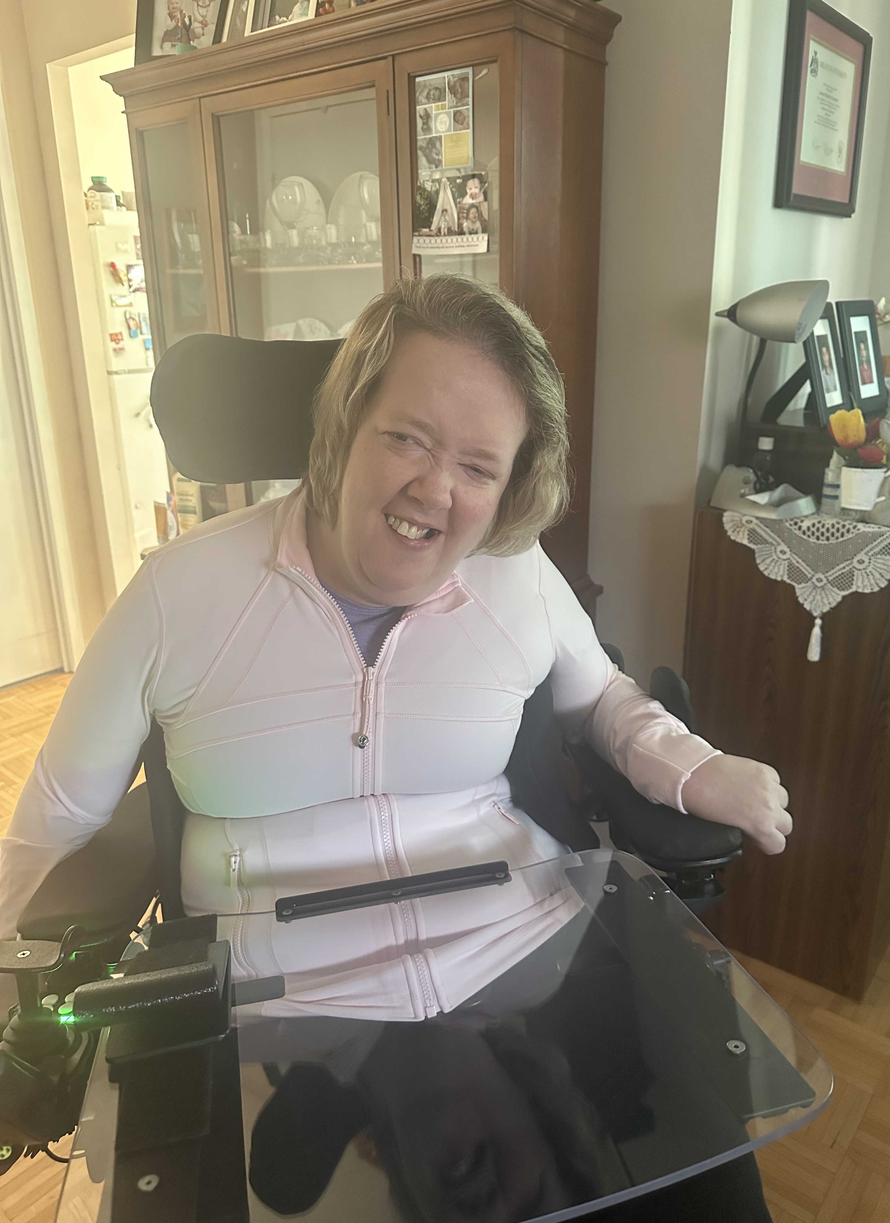 Jenny, with shoulder-length hair and wearing a light pink zip-up, smiles while sitting in a power wheelchair in a living room.