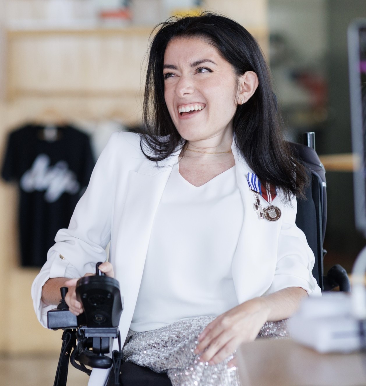 Maayan, who has long brown hair and wears a white shirt and white blazer, sits in her power wheelchair and smiles at someone out of frame. The background is blurred.