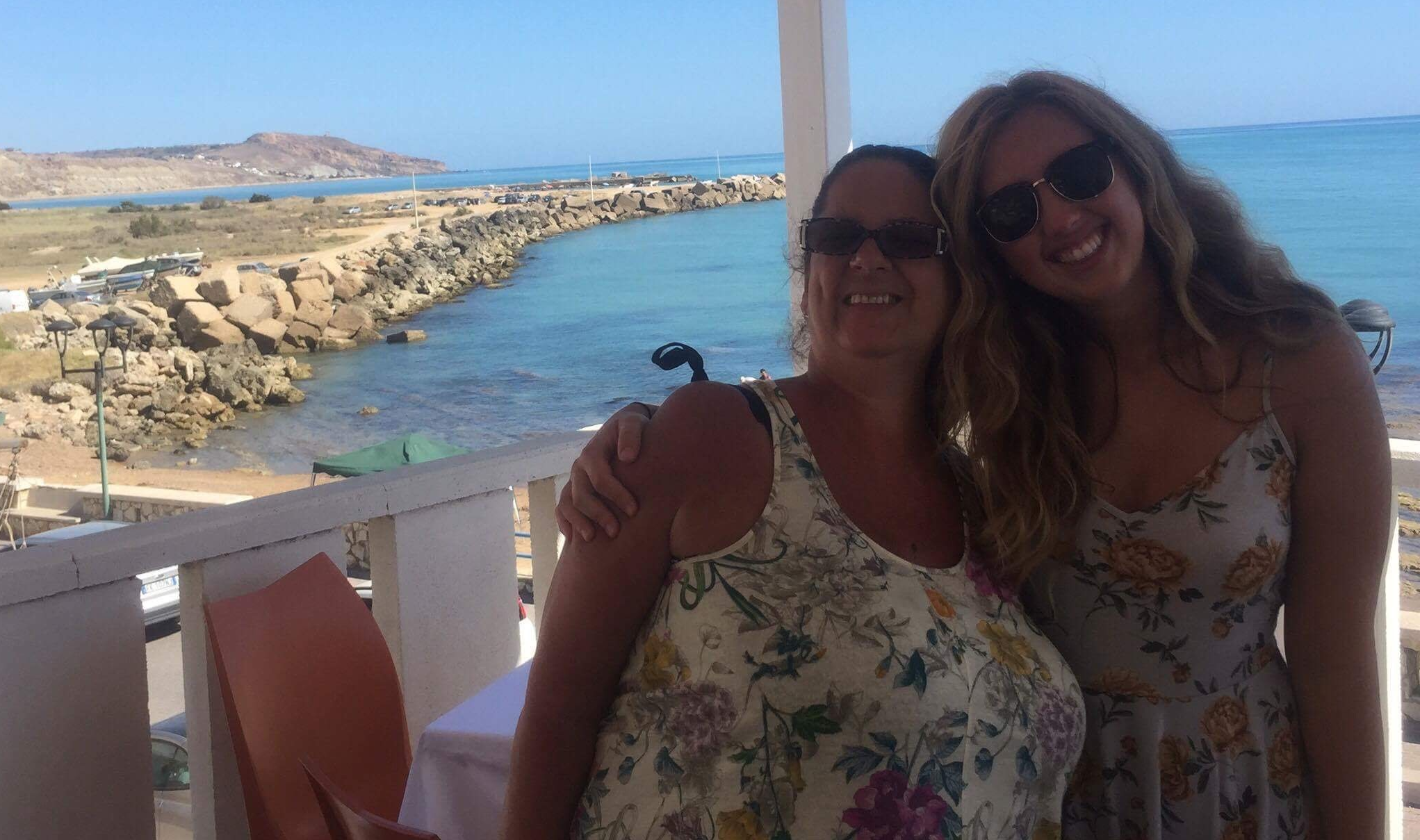 Piera and another person, both wearing white floral outfits and sunglasses, smile with their arms around each other in front of a large body of water.