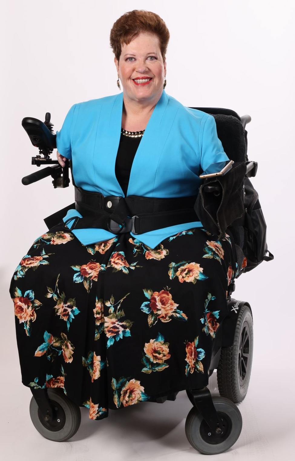 Susie, who has short brown hair and wears a light blue blazer over a black top, poses smiling in her power wheelchair against a white background.