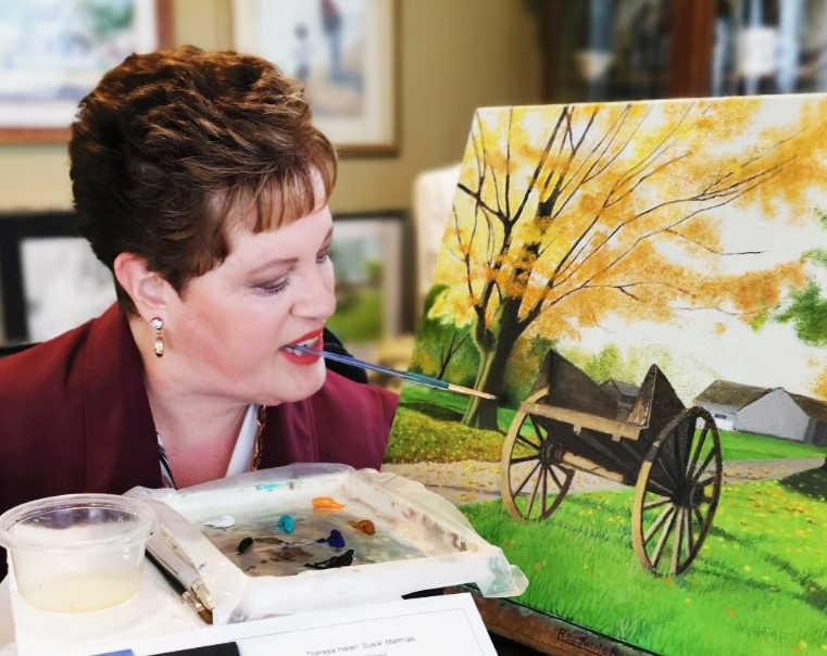 Susie paints a picture of a wagon on a grassy landscape, with a tree on one side. She holds the small paint brush between her teeth to paint. Other painting materials sit directly in front of her.